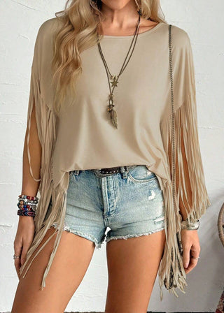 A woman wearing a beige long-sleeve top with extensive fringe detailing, styled with light-wash denim shorts, layered necklaces, and bracelets against a white textured wall.