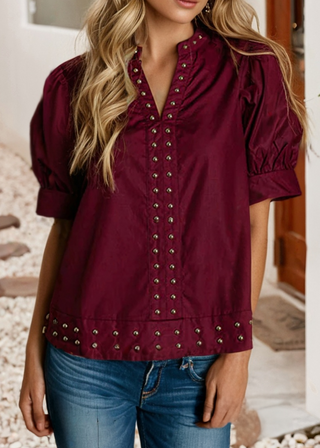 A woman wearing a burgundy short-sleeve blouse with puff sleeves and silver-tone metal studs, paired with blue jeans.
