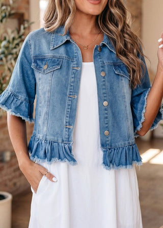 A woman wearing a light blue denim jacket with ruffled sleeves and hem over a white dress, shown in a warm indoor setting.