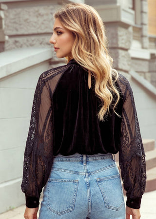 A woman shown from the back wearing a black velvet mock-neck blouse with sheer lace long sleeves, paired with light blue jeans, set against a blurred stone building background.