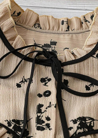 Close-up of a beige textured blouse with black floral embroidery, a ruffled collar, and a black ribbon tie at the neckline.