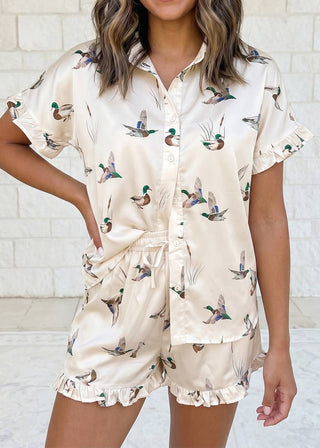 Woman wearing a beige pajama set with duck print against a white brick wall.