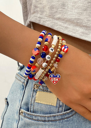 Close‑up of a wrist wearing layered red, white, blue, and gold beaded patriotic bracelets, including USA letter beads and a heart charm, styled with denim