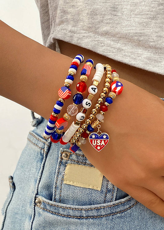 Close‑up of a wrist wearing layered red, white, blue, and gold beaded patriotic bracelets, including USA letter beads and a heart charm, styled with denim