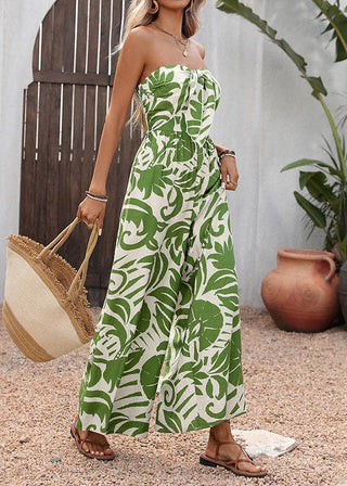 Woman wearing a strapless green leaf-patterned jumpsuit holding a straw bag outdoors.