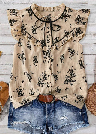 A beige sleeveless blouse with a black floral print, ruffled collar, and tie detail, styled flat-lay on a white wood background with denim shorts, sandals, a belt, and a woven bag.