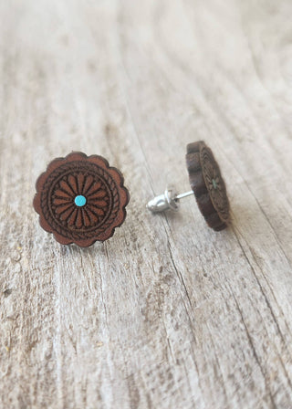 A pair of handmade wooden stud earrings with a scalloped circular design, featuring a small turquoise stone inlay at the center and a silver post, resting on a light gray wood surface.