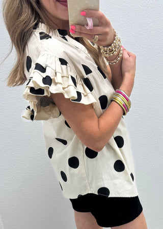 A woman taking a mirror selfie wearing a cream-colored blouse with black polka dots and ruffled sleeves, paired with black shorts.