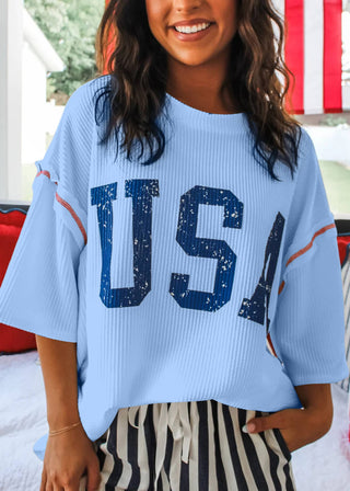 Woman wearing a blue ribbed top with bold blue “USA” lettering and red stitching