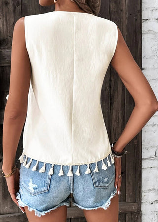 Person wearing a white sleeveless top with tassel details and denim shorts against a wooden wall.