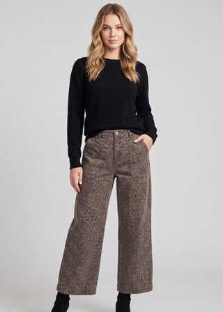A woman modeling a pair of wide-leg cropped pants with a brown and beige leopard print pattern, styled with a black long-sleeve top and black ankle boots against a light gray studio background.