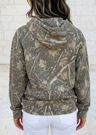 A person viewed from the back wearing a camouflage hoodie with a woodland tree-bark pattern in olive, brown, and beige, paired with white pants, standing against a light brick wall.