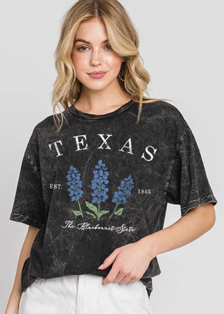 A woman modeling a black acid-wash graphic t-shirt featuring bluebonnet flowers and the word 'TEXAS' on the front, paired with white shorts against a light gray studio background.