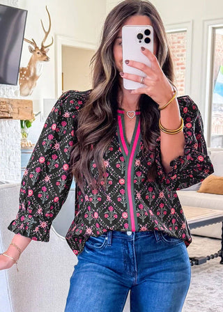 Woman taking a mirror selfie wearing a floral blouse with a pink zipper and blue jeans in a living room.