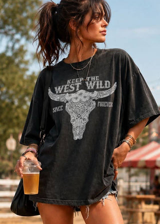 A woman stands outdoors in a rustic fairground setting wearing an oversized faded black graphic t-shirt with a white bull skull and 'KEEP THE WEST WILD' print, paired with denim shorts and red cowboy boots.