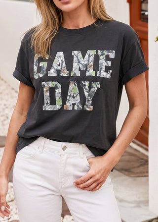 A woman wearing a dark charcoal gray graphic t-shirt with 'GAME DAY' printed in a camouflage pattern, paired with white jeans.