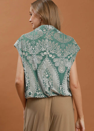 A woman shown from the back wearing a sage green sleeveless top with a white botanical paisley pattern, paired with beige pants, against a solid warm brown background.