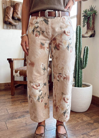 A person wearing beige wide-leg cropped pants with a vintage floral print, styled with a brown belt, brown top, and brown flats in a rustic indoor setting with a cactus and wooden floor.