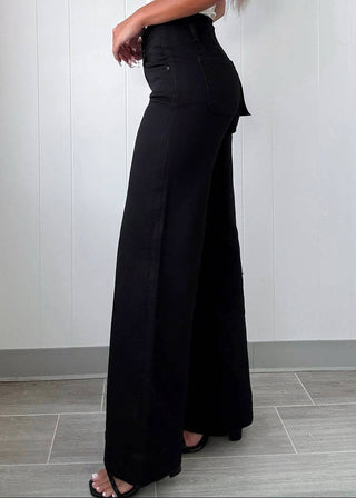 A person wearing black wide-leg pants with a draped side detail, paired with a white top and black sandals, standing against a white paneled wall.