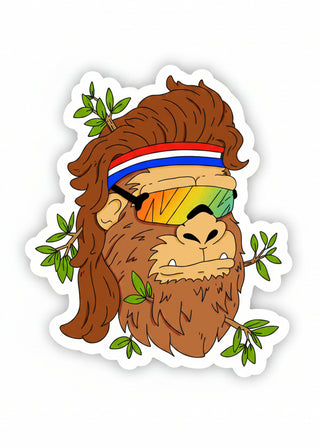 A cartoon-style die-cut sticker featuring a brown furry ape or gorilla wearing a red, white, and blue headband and rainbow-tinted sunglasses, with green leaves around the character on a white background.