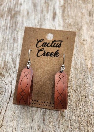 Brown leather earrings on a 'Cactus Creek' branded card against a wooden background