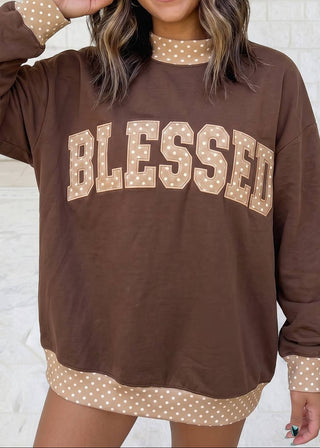 A woman wearing a brown long-sleeve sweatshirt with 'BLESSED' printed across the front in beige letters with white polka dots. The sweatshirt features a matching beige polka-dot mock neck collar, cuffs, and hem, set against a light brick wall background.