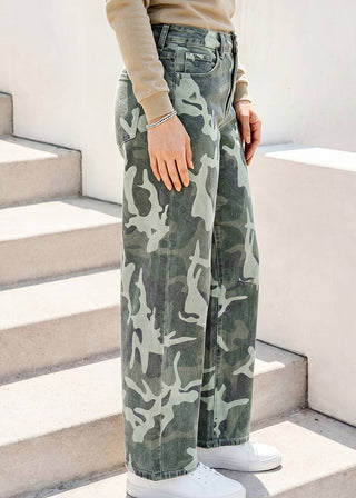 A person wearing high-waisted, wide-leg camouflage pants in green, gray, and beige, paired with a beige long-sleeve top and white sneakers, standing on concrete stairs.