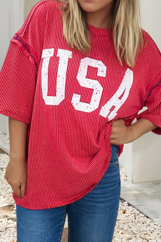 Woman wearing a red ribbed top with bold white “USA” lettering and red stitching