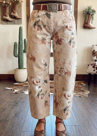 A person wearing wide-leg cropped pants with a beige base and a muted floral print in dusty pink, brown, and green, styled with a brown leather belt and brown flats in a rustic indoor setting.