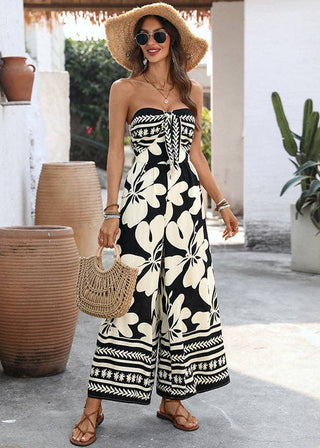 Woman in a black and white floral jumpsuit standing outdoors with plants and a white wall in the background.
