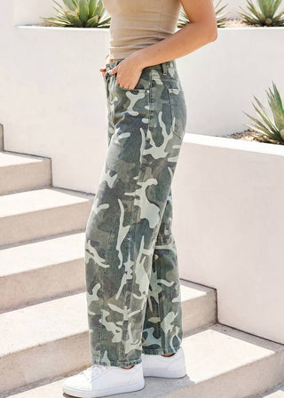 A person wearing camouflage wide-leg jeans, a beige crop top, and white sneakers, standing on outdoor concrete steps with white planter boxes and succulents in the background.