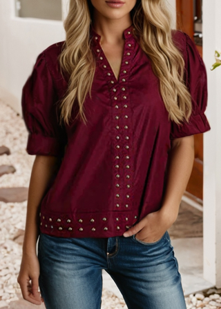 A woman wearing a burgundy short-sleeve blouse with puff sleeves and silver-tone studs, paired with blue jeans, shown in a lifestyle setting.