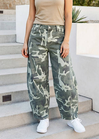 A person wearing loose-fit camouflage pants with a beige tank top and white sneakers, standing on outdoor concrete steps.