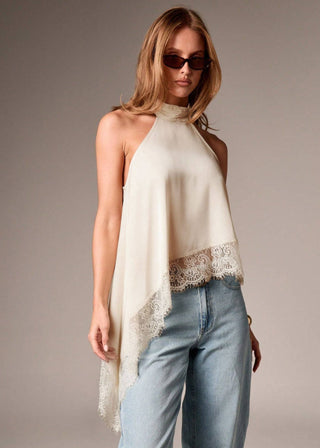 Woman wearing a beige halter top with lace trim and blue jeans against a gray background