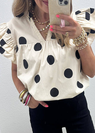 Person wearing a white blouse with black polka dots and black shorts, holding a phone.