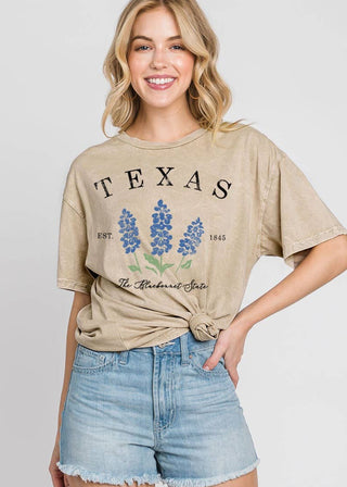 A woman modeling a beige graphic t-shirt with a distressed 'TEXAS' print and bluebonnet flowers, paired with light-wash denim shorts, against a light gray background.