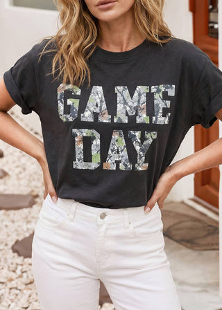 A woman wearing a black graphic t-shirt with 'GAME DAY' printed in a camouflage pattern, paired with white jeans, standing outdoors.