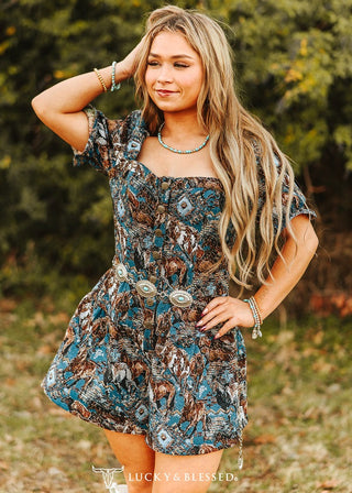 Woman wearing a blue horse‑print jacquard romper with structured puff sleeves, button‑front bodice, and turquoise accessories, styled with a silver concho belt in an outdoor setting.