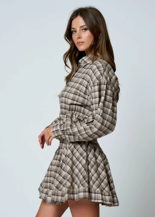 A woman modeling a long-sleeve plaid mini dress with a tie waist, shown against a plain light gray studio background.
