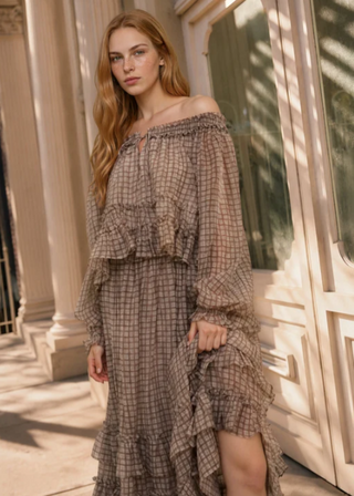 A woman models an off-the-shoulder, long-sleeve ruffled maxi dress with a brown and beige plaid pattern in an elegant outdoor setting with classical columns and warm sunlight.