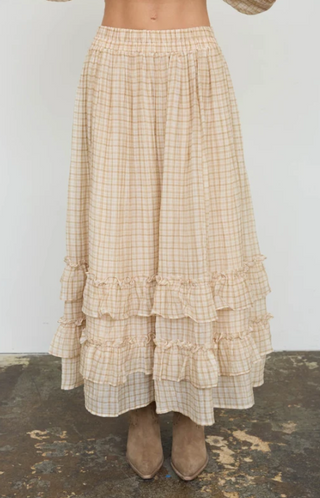 A beige and white checkered tiered maxi skirt with a ruffled waistband, worn by a model with brown suede boots against a plain white wall.