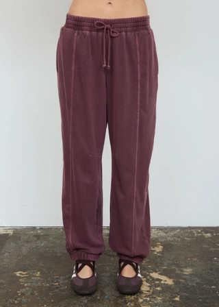 A person wearing loose, drawstring jogger-style pants in a muted plum-burgundy color, standing against a plain light wall with a worn concrete floor.