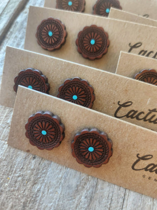 Close-up of handmade oval stud earrings with a dark brown sunburst pattern and a small turquoise center, mounted on brown kraft cards on a rustic wooden surface.