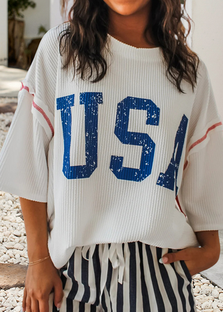 Woman wearing a white ribbed top with bold blue “USA” lettering and red stitching