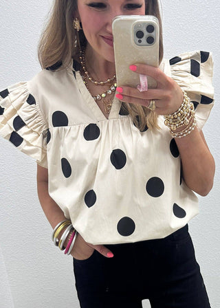 Person wearing a white blouse with black polka dots and black shorts, taking a mirror selfie.