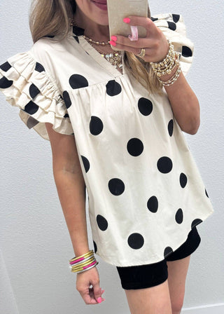 Person wearing a white blouse with black polka dots and black shorts against a light background