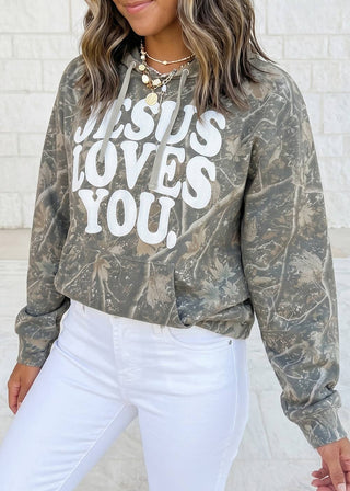 A woman wearing a long-sleeve cropped hoodie with a distressed camouflage pattern in olive green, brown, and gray, featuring the text 'JESUS LOVES YOU.' in bold white letters.