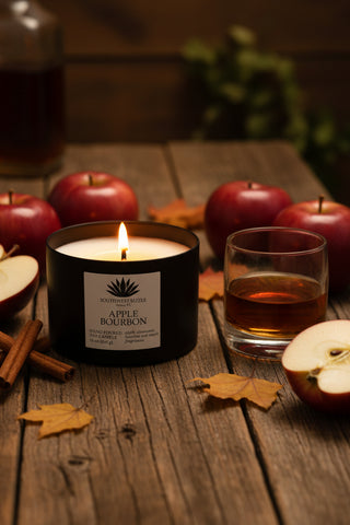 Apple Bourbon soy candle in a black tin on a wood table with apples and a glass of bourbon