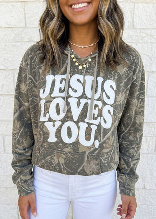 A woman wearing a cropped long-sleeve hoodie with a distressed camouflage pattern in olive green and brown, featuring the phrase 'JESUS LOVES YOU.' printed in large white letters across the front.
