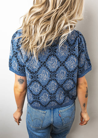 Person wearing a blue patterned top and jeans with a plain background